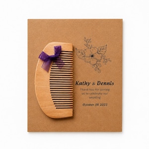 Può includere: Un pettine in legno marrone chiaro decorato con un nastro viola, fissato su un cartoncino beige con un disegno floreale nero e il testo: Kathy & Dennis, Thank You for joining us to celebrate our wedding, October 08 2023.