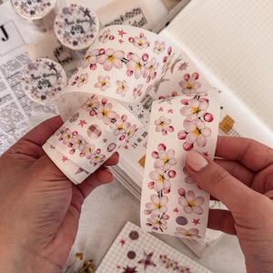 Può includere: Un rotolo di nastro adesivo decorativo con un motivo ripetuto di fiori di ciliegio rosa e bianchi, piccole stelle rosa e motivi a quadri circolari su sfondo color crema. Il nastro viene srotolato per mostrare il disegno floreale.