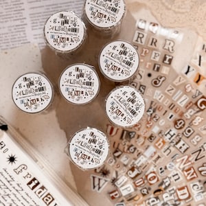 Könnte beinhalten: Eine Sammlung von Washi-Tape-Rollen und Buchstabenaufklebern. Die Tape-Rollen sind weiß mit schwarzem Text und Sternen. Die Aufkleber sind transparent mit schwarzen und braunen Buchstaben.