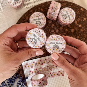 Puede incluir: Varios rollos de cinta washi con un patrón repetitivo de flores de manzano rosas y blancas. Cada rollo tiene una etiqueta circular con el texto Apple Blossom Buffy. La cinta se muestra en formato de rollo y como una tira sobre papel cuadriculado.