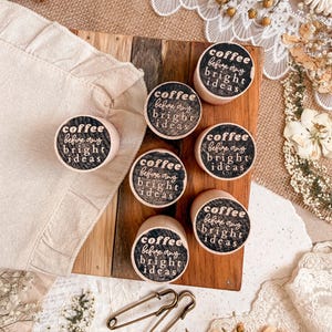 May include: A collection of small, round wooden objects with the text "coffee before any bright ideas" printed on them. The objects are arranged on a wooden board and a beige cloth. Two safety pins are also visible.