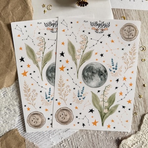 Könnte beinhalten: Zwei Stickerbögen mit himmlischen und botanischen Designs. Jeder Bogen enthält einen Mond, Sternbilder, Sterne und florale Elemente. Die Aufkleber haben einen Aquarellstil mit orangefarbenen, grünen und schwarzen Akzenten. Der Text "#629 Bujo Jej" ist sichtbar.