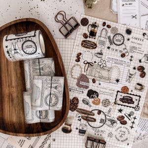 May include: A collection of coffee-themed stationery items including rolls of decorative tape with coffee motifs, a grid-patterned sticker sheet featuring various coffee illustrations and text, and two metal binder clips on a wooden tray.