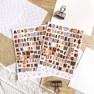Op de afbeelding: Twee stickervellen met alfabetletters uit verschillende papiertexturen, in een collagestijl. De letters zijn in verschillende lettertypen en kleuren, waaronder oranje, bruin en groen. De vellen liggen op een achtergrond van vintage papier en kant.