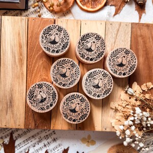 Könnte beinhalten: Sechs runde Holzstempel mit einer schwarzen Tintenillustration eines Vogels, der zwischen Blättern und Eicheln sitzt. Jeder Stempel hat eine natürliche helle Holzmaserung und eine flache Oberseite mit dem detaillierten botanischen Design.