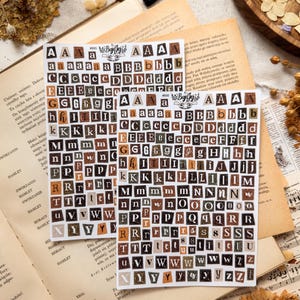 Op de afbeelding: Twee vellen met alfabetstickers met individuele letters in verschillende lettertypes en groottes, gedrukt op kleine rechthoekige stukjes papier in bruin-, beige-, zwart- en crèmetinten. Elk vel bevat bovenaan de merknaam BujoJej.