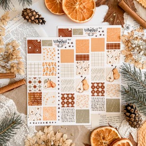 Pode incluir: Duas folhas de autocolantes decorativos com padrões de peras, estrelas e xadrez em tons de castanho, laranja e bege. Rodelas de laranja secas, pinhas e paus de canela são dispostos ao redor dos autocolantes, criando um tema de outono.
