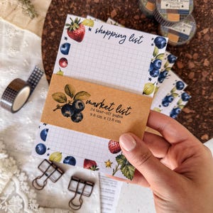 Può includere: Un blocco note rettangolare a griglia, decorato con illustrazioni di fragole, mirtilli e uva spina lungo i bordi. Il testo riporta shopping list, market list, 24 tear-off pages, 9.8 cm x 13.8 cm.
