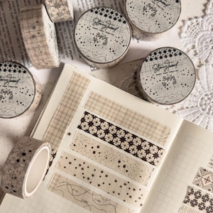 Puede incluir: Un surtido de rollos y muestras de cinta washi decorativa con varios diseños, incluyendo cuadros, lunares y diseños geométricos. Los rollos de cinta presentan el texto "Story Trend BujoJej". La paleta de colores es neutra, con tonos beige, marrón y negro.