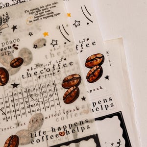 May include: Overhead shot of coffee-themed decorative stickers. The stickers feature coffee beans, stars, and text like "the coffee" and "life happens coffee helps." The stickers are on a translucent background.