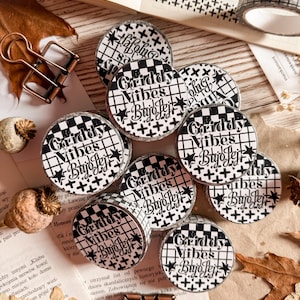 Puede incluir: Varios rollos de cinta washi con un patrón de cuadrícula en blanco y negro y el texto "Griddy Vibes Bujo Jej". La cinta está sobre una superficie de madera con papelería y hojas secas.