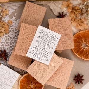 Puede incluir: Una colección de pequeñas tarjetas rectangulares de madera con escritura impresa, algunas apiladas y otras dispersas. Una pequeña tarjeta blanca con texto manuscrito encima. Rodajas de naranja secas, anís estrellado y encaje añaden a la estética vintage.