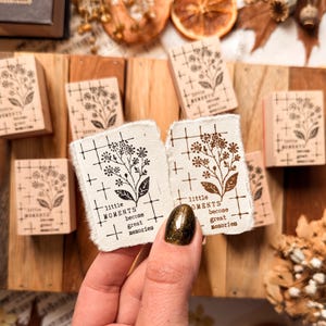 Op de afbeelding: Twee rechthoekige, gebroken witte stukjes papier met gescheurde randen, elk voorzien van een zwarte inktstempel van een bloemtakje, een rasterpatroon en de tekst "little MOMENTS become great memories" in een verticale opstelling.