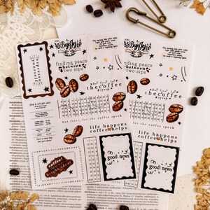 May include: A collection of coffee-themed stickers with various designs and text, including coffee beans, stars, and phrases like "finding peace between two sips" and "life happens coffee helps." Also, two antique brass safety pins and coffee beans.
