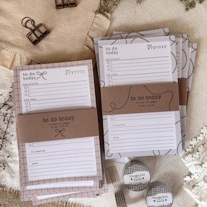 May include: Two stacks of to-do list notepads with 25 tear-off pages each, measuring 12 cm x 18 cm. One stack features a grid pattern, the other has abstract black lines. Both are wrapped in brown paper bands labelled 'to do today'.