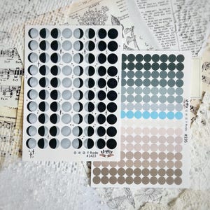 May include: Two sticker sheets with celestial and dot designs. One sheet features moon phases in black, grey, and white, with star accents. The other has gradient dots in shades of grey, blue, and brown. The stickers are on a white background with vintage paper.