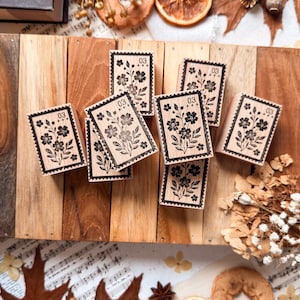 Puede incluir: Una colección de sellos de madera rectangulares con una ilustración en tinta negra de un ramo de flores con el número 03 y tres signos más impresos en la parte superior. Cada sello tiene un borde dentado que recuerda a un sello postal.