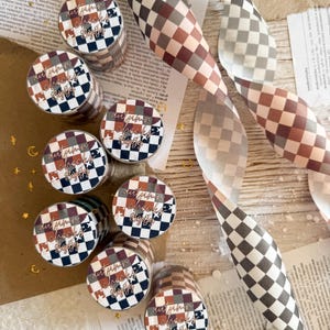 Pode incluir: Uma coleção de rolos de fita adesiva decorativa e fita com um padrão xadrez em tons de castanho, branco e preto. Os rolos de fita adesiva têm o texto "autumn checks byjojo". A fita é enrolada e colocada sobre uma superfície texturizada.