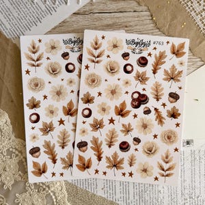 Può includere: Due fogli di adesivi con disegni a tema autunnale. Gli adesivi includono illustrazioni di foglie, fiori, ghiande e stelle nei toni del marrone, beige e crema. Il testo "Bujo Jej" e "#763" è visibile.