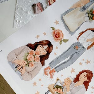 A close-up of the sticker sheet, showing detailed watercolor illustrations of women with soft expressions holding peach-colored roses. Surrounding them are tiny stars and floral stickers in warm, muted tones.