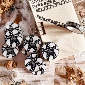 May include: A collection of Halloween-themed washi tape rolls and decorative tape strips. The rolls feature a black and white ghost design with the text "checker BOO BujoJej". The tape strips have checkerboard, ghost, and pumpkin patterns.