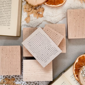 Op de afbeelding: Een verzameling lichtbruine houten stempels met een rasterpatroon. Sommige stempels zijn opgestapeld, andere zijn gerangschikt op een oppervlak met open boeken, gedroogde sinaasappelschijfjes en kant.