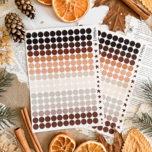 May include: Two sheets of dot stickers in a gradient of neutral colours, from black to light beige. The stickers are arranged in rows of circles. The image also includes dried orange slices, pine cones, and cinnamon sticks.