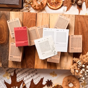 May include: A collection of rectangular wooden stamps featuring grid patterns for daily planning, each with a space for a date and notes. One stamp has a red rubber face, while others are natural wood. Several stamped paper samples are included.