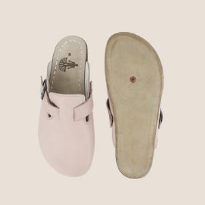 May include: Two light pink clogs, one showing the top and the other the sole. The top clog has a buckle strap and a leather insole with a tree logo and the number 40. The sole is light brown with a copper-coloured rivet.