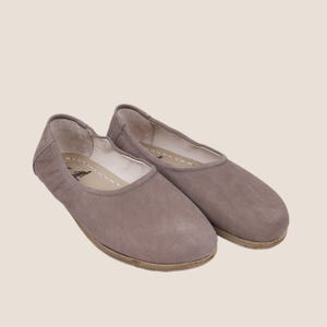 May include: A pair of taupe-colored ballet flats. These slip-on shoes have a rounded toe and a flat sole. The upper is made of a soft, suede-like material. The interior is lined with a lighter-colored material, and the sole is a light brown.