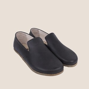 May include: A pair of black leather slip-on shoes. The shoes have a rounded toe and a low profile. The soles are a light brown color. The shoes are designed for comfort and casual wear.