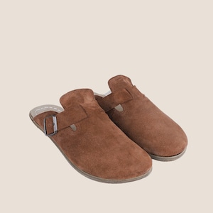 May include: A pair of brown suede clogs. Each clog features a buckle closure and a rounded toe. The clogs have a slip-on design and a neutral-colored sole. The shoes are made of a soft, textured material.