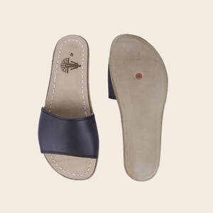 May include: Black leather slide sandals with a natural-colored footbed and white stitching. The sandals have a simple design with a single strap across the top of the foot. The sole is a light brown color. The sandals are size 40.