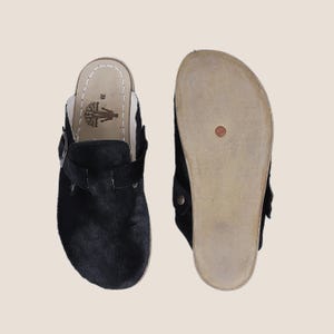 Può includere: Due zoccoli in pelle nera, uno che mostra la parte superiore con un cinturino con fibbia e l'altro che mostra la suola. Gli zoccoli hanno una soletta color naturale e un esterno nero strutturato. La suola è color beige chiaro.