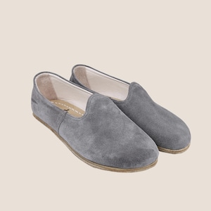 May include: A pair of gray suede slip-on shoes. The shoes have a rounded toe and a low profile. The soles are a light brown color. The interior of the shoe is a light beige color.