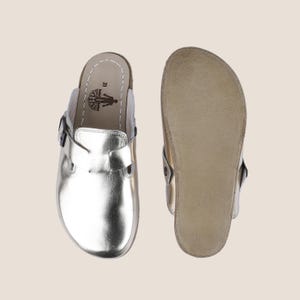 May include: Metallic silver clogs with a buckle closure. The clogs have a cork footbed and a leather upper. The sole is a light brown color. The clogs are shown next to their sole.
