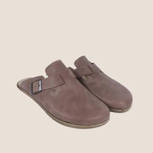 May include: A pair of taupe leather clogs with a buckle fastening. The clogs have a rounded toe and a strap across the top. The soles are a lighter brown colour. The clogs are set against a neutral background.