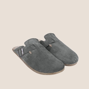May include: A pair of gray suede slip-on clogs. The clogs have a buckle closure and a light brown sole. The shoes are designed for comfort and casual wear.