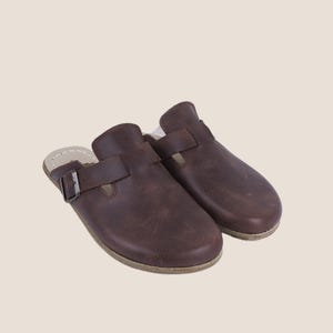 Może przedstawiać: Para brązowych skórzanych chodaków z zapięciem na klamrę. Chodaki mają zaokrąglony nosek i pasek na górze. Podeszwy są w jasnobrązowym kolorze. Chodaki są przeznaczone do noszenia jako buty wsuwane.