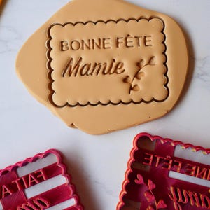 Peut inclure: Deux emportes-pièces en plastique rouge en forme de rectangle avec les mots "Bonne Fête Mamie" et une branche de feuilles. Les emportes-pièces sont sur une surface en marbre blanc.