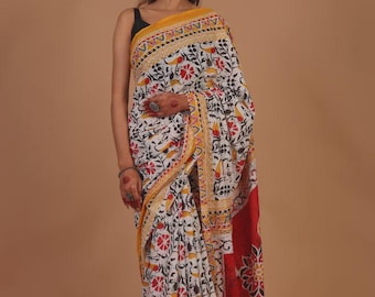 May include: A white saree with a floral and bird pattern in red, black, and yellow. The saree has a yellow border and a red panel with a large floral design. The woman is wearing a black top.