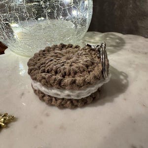May include: A handmade crochet keychain resembling a sandwich cookie. The cookie has a brown exterior and a white filling. A silver-colored keyring is attached to the top. The background includes a decorative glass sphere and a marble surface.