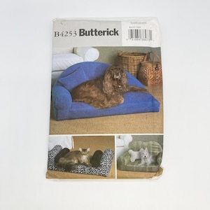May include: A Butterick B4253 sewing pattern envelope featuring illustrations of pet beds. The top shows a blue sofa-style bed with a brown dog. The bottom left shows a black and white leopard print bed with a cat, and the bottom right shows a green bed with a small dog.