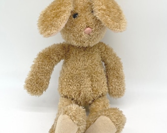 May include: A plush stuffed animal in the shape of a rabbit with light brown, shaggy fur. The rabbit features long, floppy ears, dark round eyes, and a small, light pink stitched nose. The feet have smooth, light beige fabric pads on the soles.