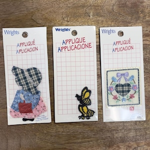 May include: Three Wrights brand iron-on appliques on white cards. Designs include a gingham girl, a yellow and black butterfly, and a heart-shaped wreath with floral accents. Each card has the words "APPLIQUÉ APLICACION" printed on it.