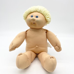 May include: A vintage Cabbage Patch Kid doll with a soft, tan body and a yarn hairstyle. The doll has blue eyes and a smiling expression. It is sitting with its arms and legs outstretched.