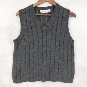 May include: A gray, sleeveless sweater vest with a V-neck design. The vest has vertical ribbed detailing and is hanging on a wooden hanger. The label reads "Malcolm Stone".