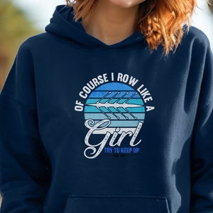 Puede incluir: Sudadera con capucha azul marino con un gráfico frontal que dice "Of course I row like a Girl, Try to keep up". El gráfico presenta un diseño de barco de remo en tonos azules y blancos. La sudadera tiene un bolsillo delantero y una capucha.