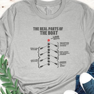 May include: A gray t-shirt with a humorous graphic depicting the different parts of a rowing boat. The text reads "The Real Parts of the Boat" with a diagram showing "Loud Mouth", "Smarties", "Complainers", and "Jokers".