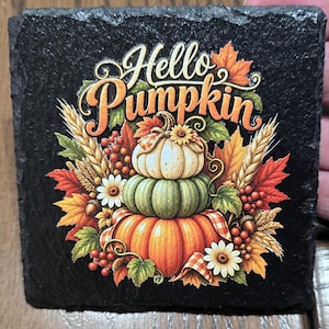 Puede incluir: Un posavasos cuadrado de pizarra negra con un gráfico colorido de tres calabazas apiladas en blanco, verde y naranja. El diseño incluye hojas de otoño, tallos de trigo, bayas rojas, flores blancas y el texto Hello Pumpkin en color naranja.